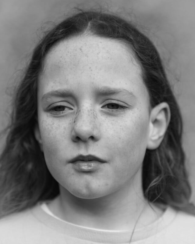 Black and white close-up portrait of a young person with long curly hair, freckles, and a neutral facial expression, looking slightly away from the camera. The background is softly blurred.