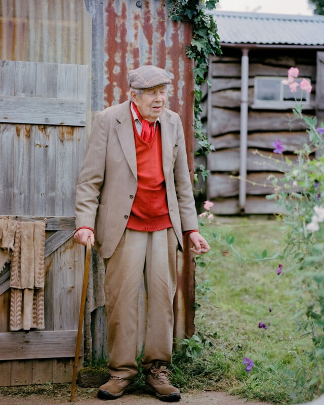 An elderly man in a flat cap, tan suit, and red sweater stands outside by a rustic wooden shed, holding a cane and surrounded by green plants and blooming flowers.