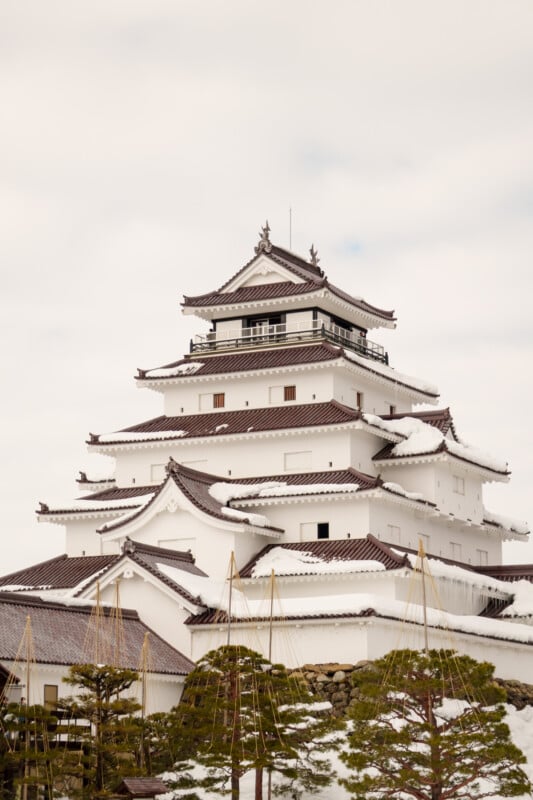 The image shows a traditional Japanese castle with multiple tiers and white exterior. The rooftops are dusted with snow. Slender trees are in the foreground, and the sky is overcast.
