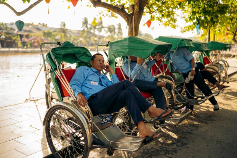 Three rickshaw drivers in blue shirts relax on their vehicles by a riverside shaded by trees, with sunlight filtering through the leaves and peaceful surroundings in the background.
