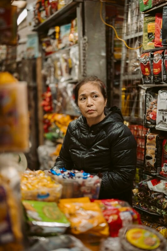 A woman wearing a black puffer jacket stands behind the counter of a small, crowded store filled with various colorful packaged snacks and goods. She looks thoughtfully to the side.