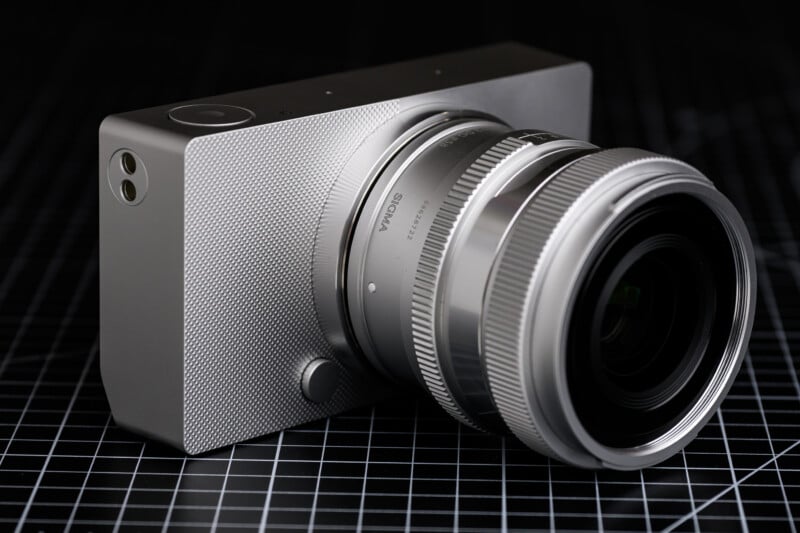 A silver digital camera with a textured body and prominent lens sits on a black grid-patterned surface. The design appears modern and sleek, highlighting the camera's compact and angular shape.