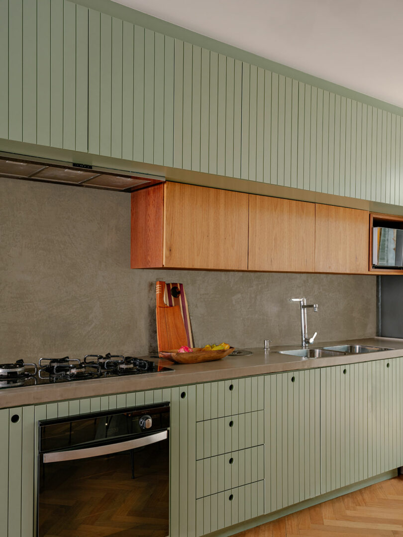 Modern kitchen with green vertical panel cabinets, wooden upper cabinets, stainless steel sink, gas stove, and a gray backsplash. A cutting board and fruit bowl sit on the counter.
