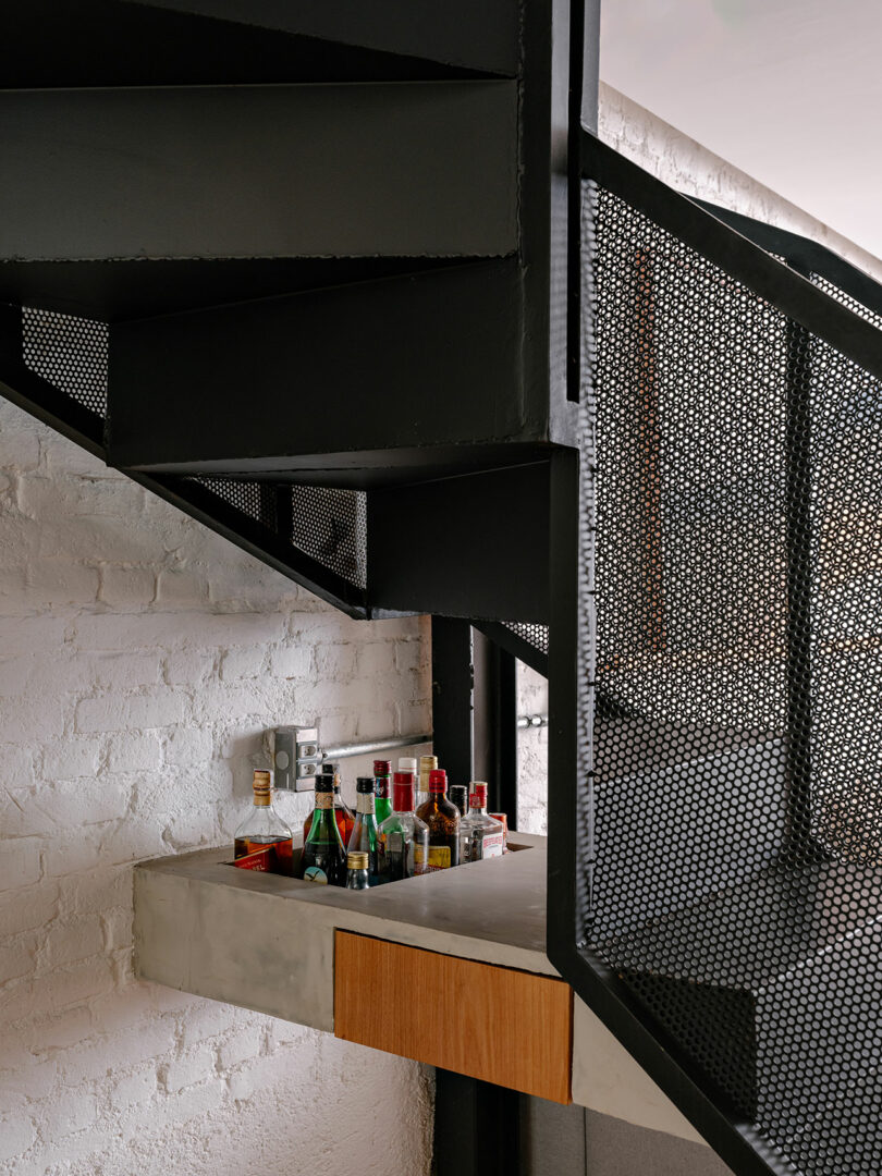 A small bar setup with assorted bottles is placed under a modern black metal staircase with a perforated railing and white brick wall backdrop.