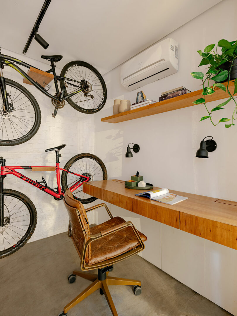 A modern home office with a wooden desk, brown swivel chair, wall-mounted shelves, two bikes on the wall, plants, books, and wall lights.