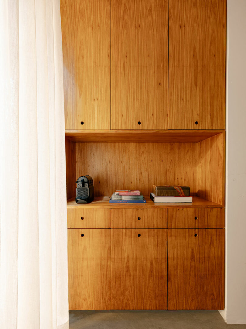 Wooden built-in cabinet with closed doors, an open shelf displaying a coffee machine, stacked books, and natural light coming from sheer curtains on the left.