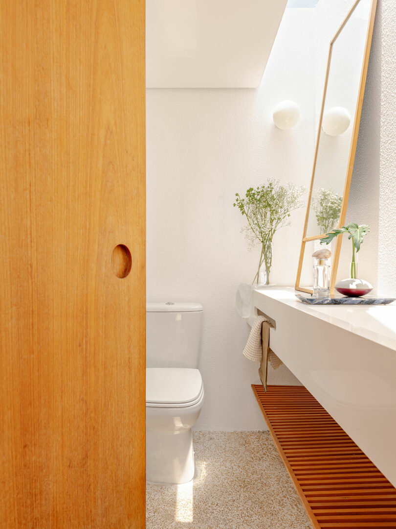 A modern bathroom with a wooden sliding door, a white toilet, a long white sink, a large mirror, and vases with greenery on the countertop. Natural light enters from above.