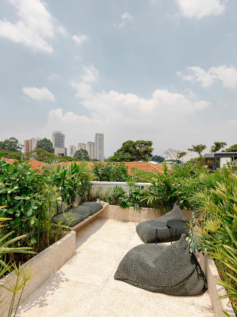 A rooftop terrace with green plants, two woven lounge chairs, a small black table, and city buildings visible in the background under a partly cloudy sky.