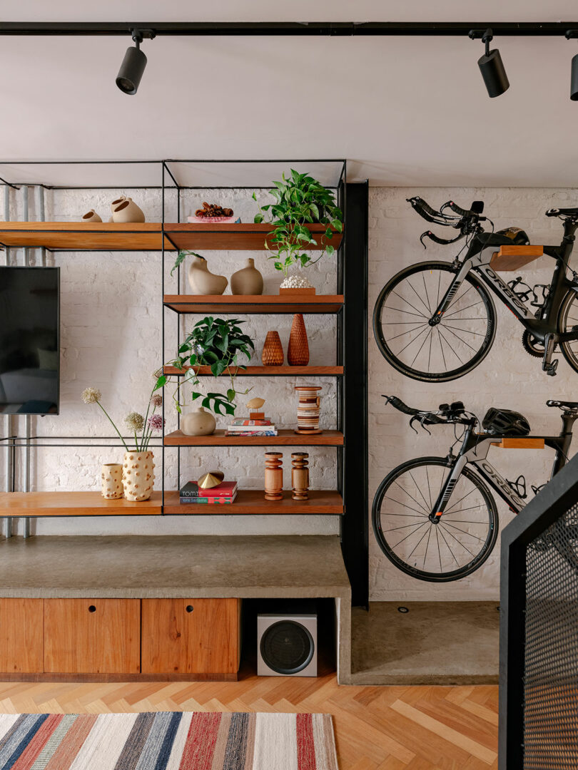 Modern living room with open wooden shelves displaying decorative items, two bicycles mounted on the white brick wall, a TV, and a striped rug on a herringbone wood floor.