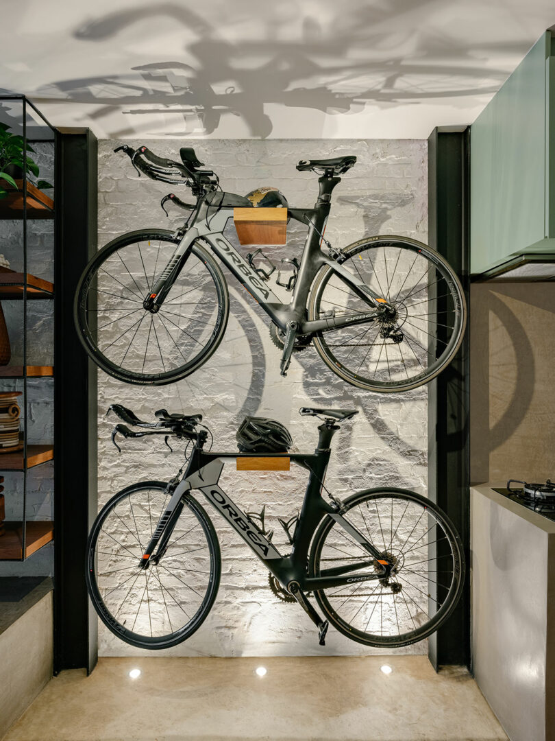 Two Orbea road bikes mounted vertically on a white brick wall, each with a wooden shelf holding cycling accessories between them, in a modern indoor space.