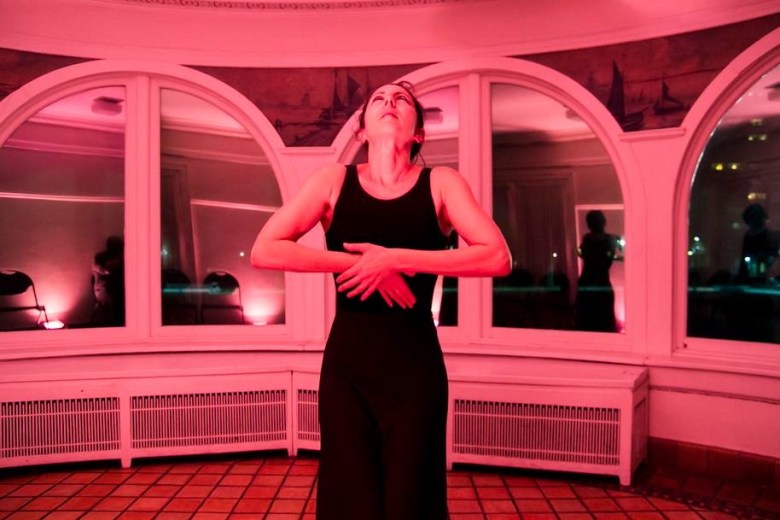 Deborah Goodman wears a black leotard and trousers, standing looking upward with her hands crossed across her torso and elbows akimbo. She is in a room with a red tiled floow and a series of arched windows with murals of ships between them. Old white radiators are underneath the windows