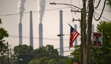White-gray emissions billow from the stacks in the background. In the foreground is an American flag on a telephone pole.