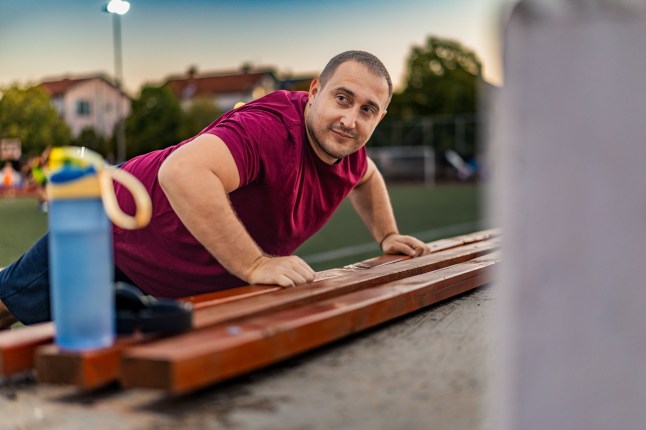 Overweight man exercising outside, doing push-ups on a bench next to the sports field.