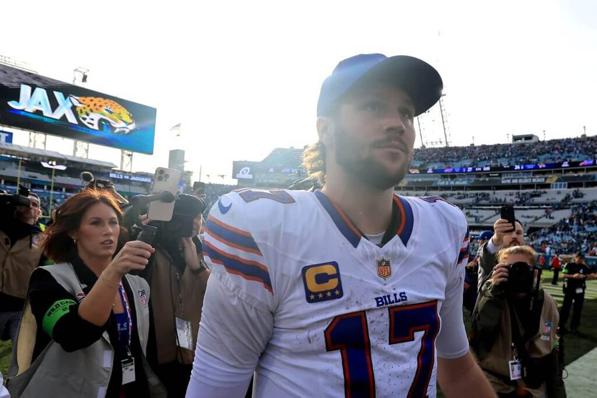 A quarterback is followed by media after a win while sunlight peaks over his right shoulder.