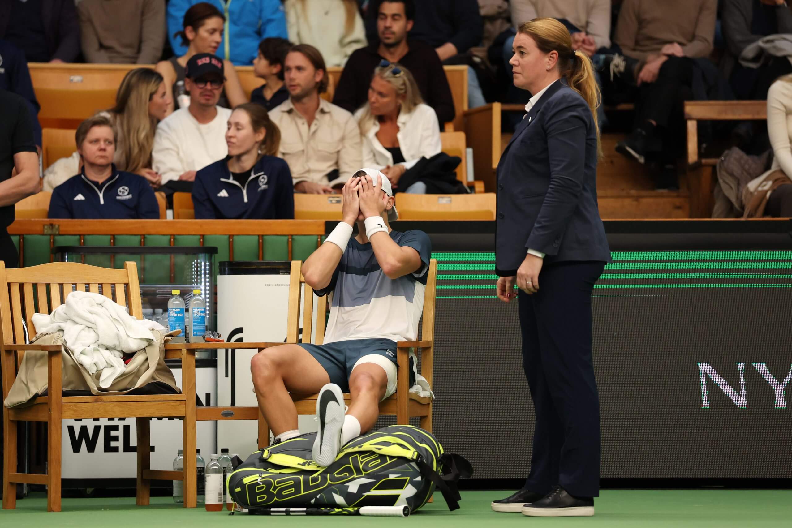Holger Rune sits in a chair with his head in his hands, with his left foot shoeless and supported by a tennis bag, as a chair umpire stands next to him.