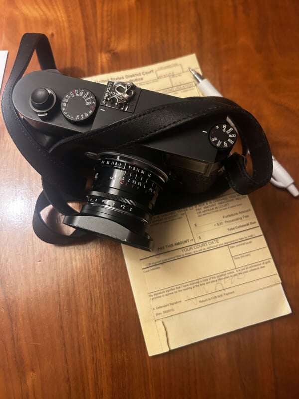 A black camera with a lens and strap rests on top of a yellow parking citation and a silver pen on a wooden surface.