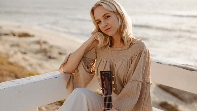 Seated woman holding acoustic guitar on wooden deck overlooking the beach and ocean.