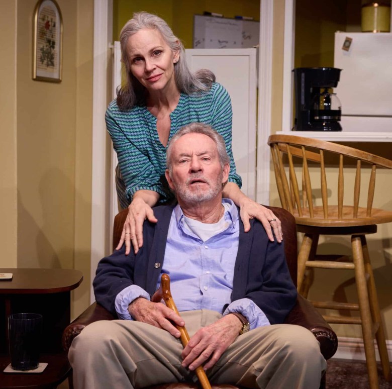 A woman stands with her hands on the shoulders of a seated old man with a cane