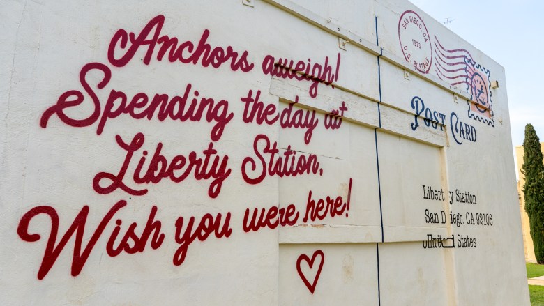 The sign is styled like a letter and reads "Anchors aweigh! Spending the day at Liberty Station, Wish you were here!"