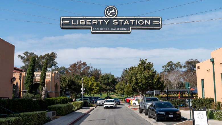 A sign reading "Liberty Station San Diego, California" hangs in the air, suspended by wires from buildings to its left and right.