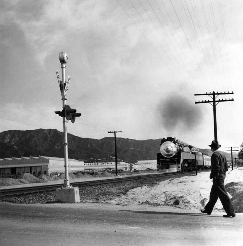 A man in a hat crosses railroad tracks as a steam train approaches in the distance, with mountains and industrial buildings in the background under a cloudy sky.