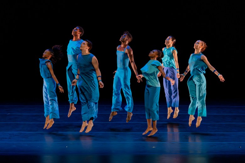 Seven dancers of varying genders and races are show midleap on stage. They are all wearing clothing in shades of blue or blue-green, they are looking slightly upward, and their arms are slightly extended at their sides.