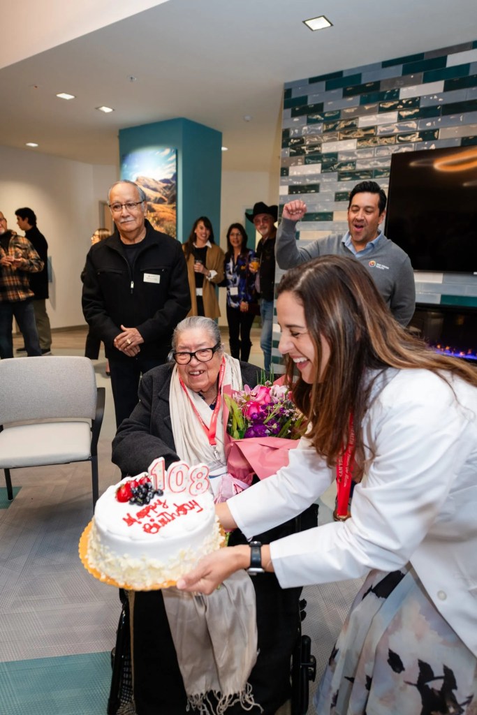 An elderly person celebrating a 108th birthday with a cake and flowers, surrounded by smiling people.
