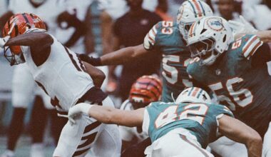 Football players in teal and orange uniforms tackle a player in a white and orange uniform during a game, with intense action and movement visible on the field.