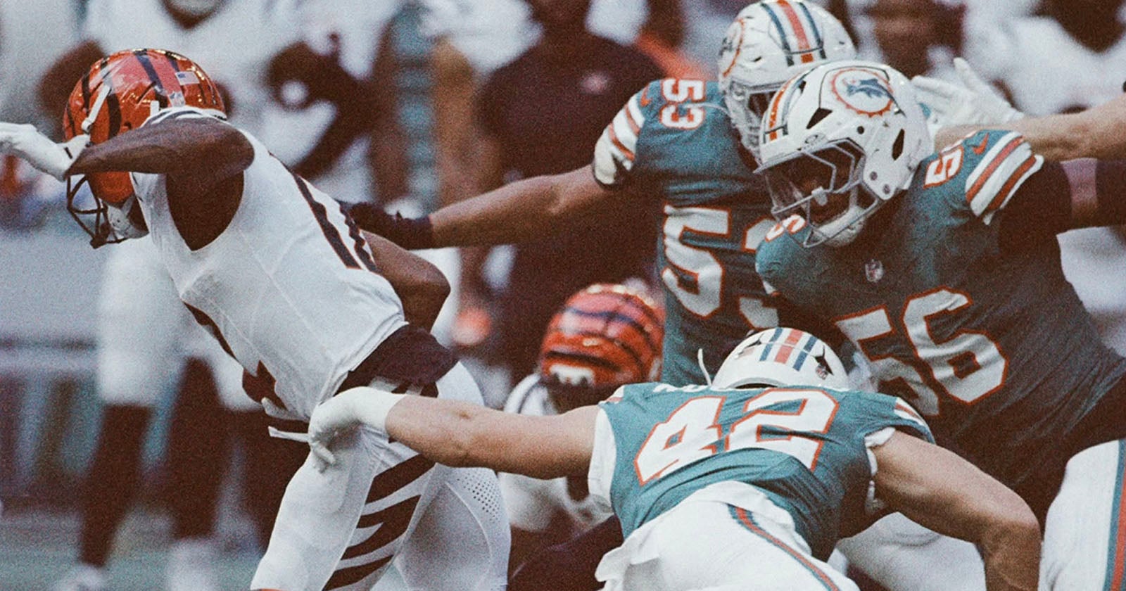 Football players in teal and orange uniforms tackle a player in a white and orange uniform during a game, with intense action and movement visible on the field.