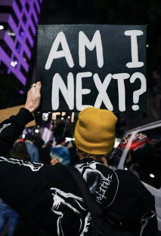 A person wearing a beanie holds up a black sign, painted in white that reads "Am I next?"