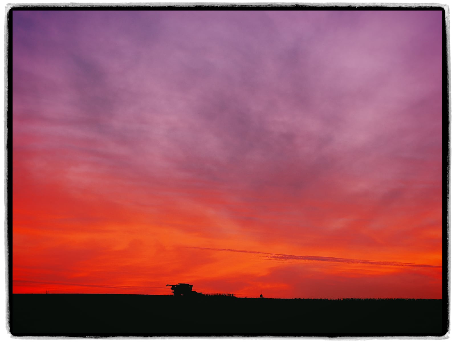 A combine harvester is silhouetted against a vibrant sunset sky filled with shades of orange, red, and purple over a flat landscape.
