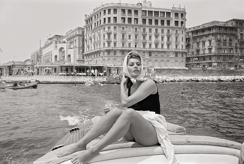 Sophia Loren sits on the deck of a boat in a black swimsuit and white headscarf, with a large grand hotel and a city coastline in the background.