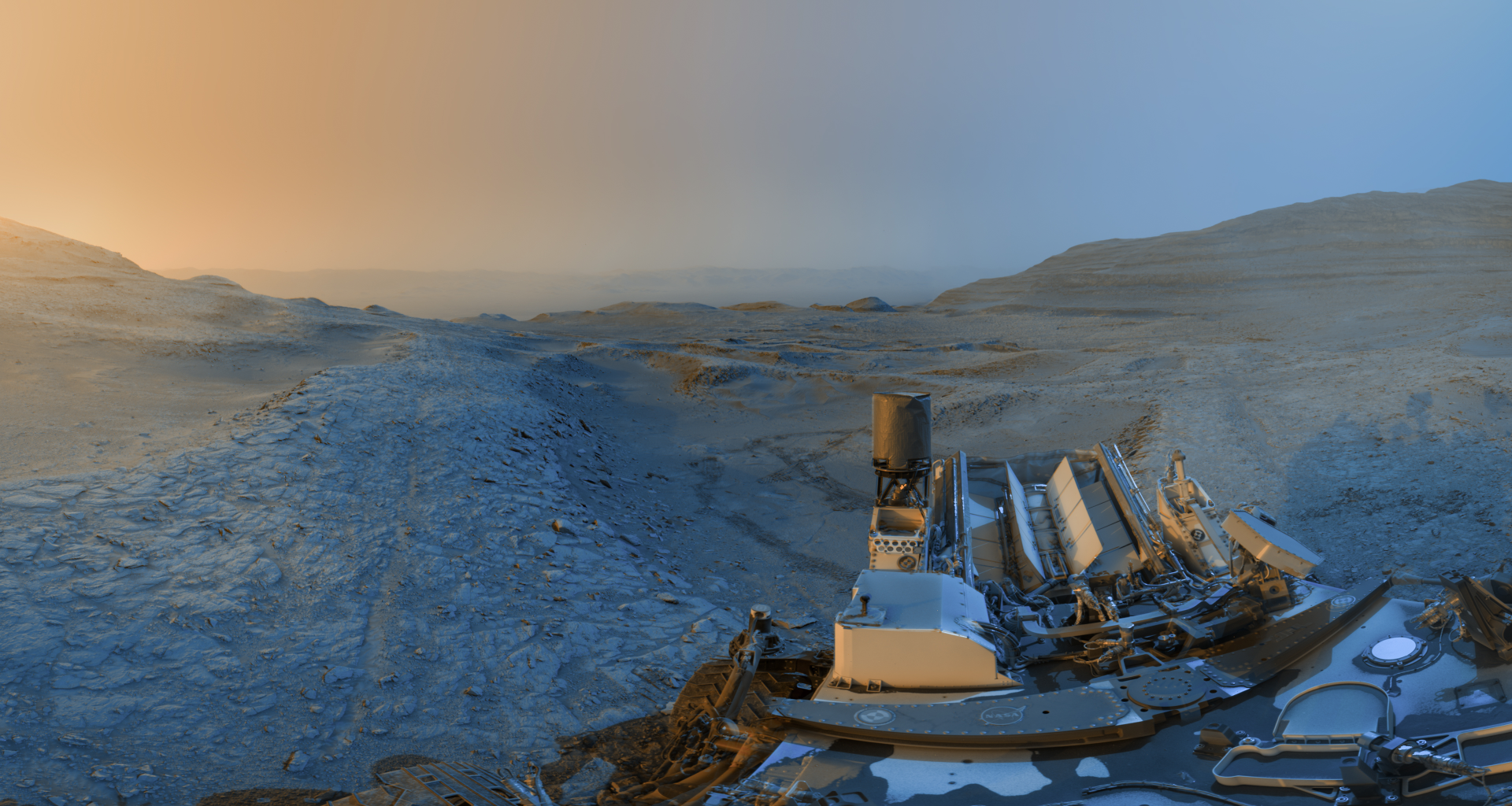 A time lapse panorama of the Martian landscape, showing blue/gray dirt with a sunset to the left and a close up of Curiosity in the bottom right of the image