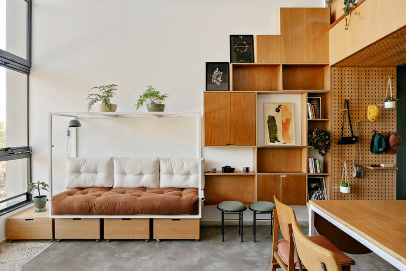 Modern living space with a cushioned sofa, wooden modular shelves, artwork, plants, and a table with chairs; natural light streams in through large windows.