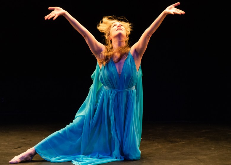 Sandra Kaufmann in a flowing sky0blue grecian gown. She is on one knee, with the other leg stretched to her side, and her arms and face upraised.