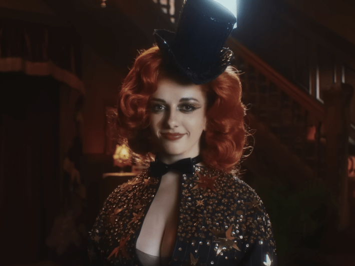 a woman in a top hat and sparkly cardigan smiles into the camera