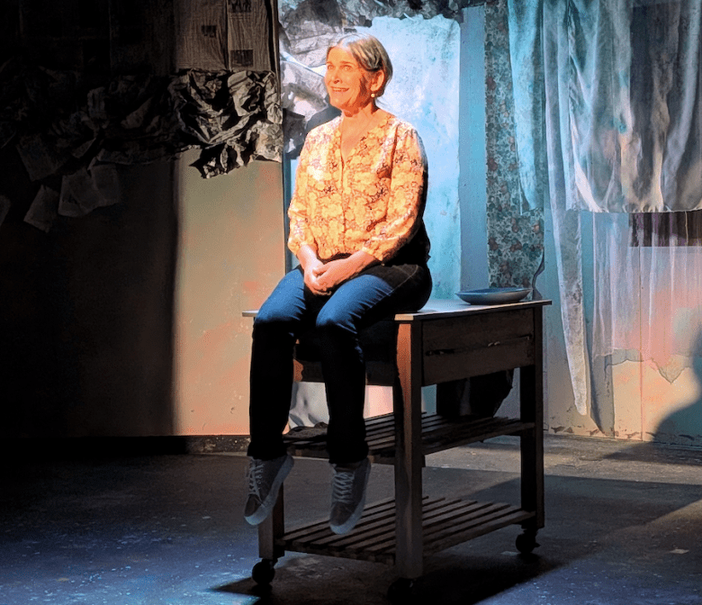 Woman seated on a rolling cart under a spotlight on a stage.