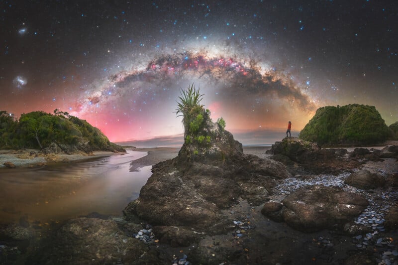 A person stands on rocky terrain near water under a starry night sky with the Milky Way arcing brightly overhead; lush green plants and cliffs frame the scenic landscape.