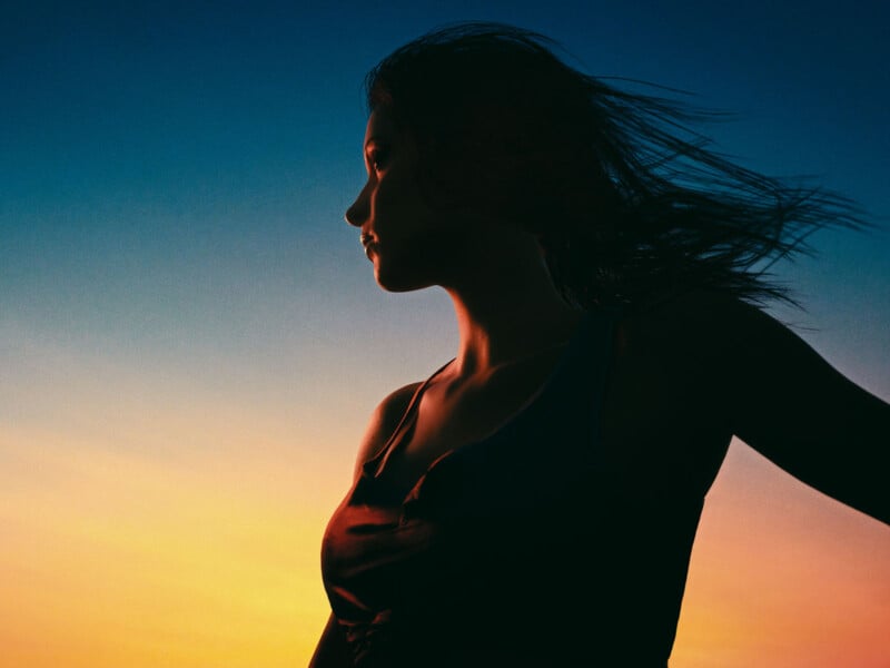Silhouette of a woman with long hair blowing in the wind, set against a vibrant sunset sky with blue, orange, and yellow hues.