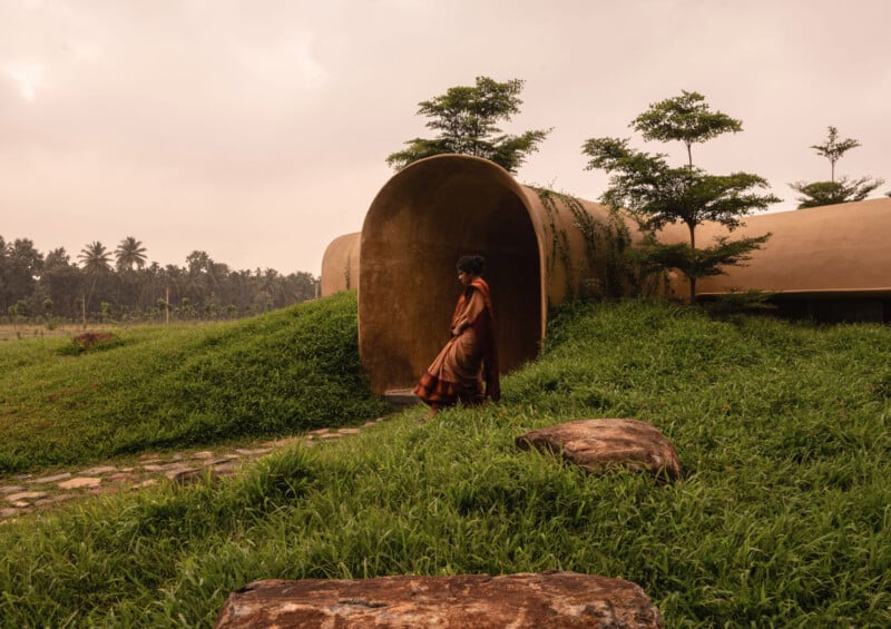 A person in traditional attire stands at the entrance of a cylindrical, earth-toned building set in grassy, rolling hills with small trees and a cloudy sky in the background.
