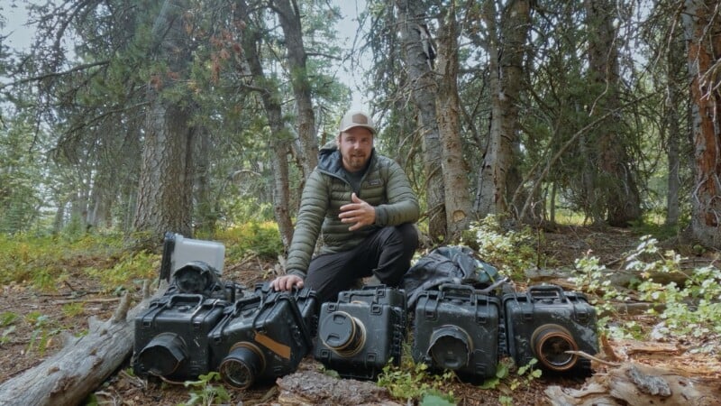 A man in outdoor clothing sits in a forest clearing, surrounded by several rugged, black hard cases. Sunlight filters through the tall trees in the background. He appears to be talking or gesturing.