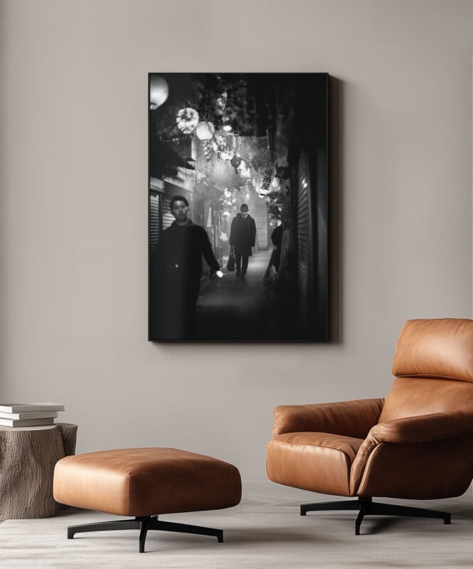 A modern living room with a tan leather armchair and ottoman, a small wooden side table with a stack of books, and a large black-and-white photograph of people walking down a lantern-lit street hanging on the wall.