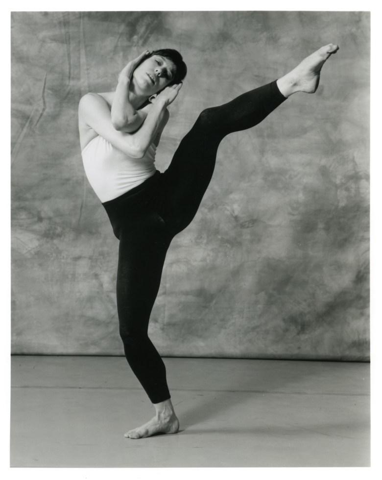 Winifred Haun is shown in a black and white photo, wearing a white tank top and black leggings. Her arms are entwined around her head, which is bent to one side. She stands on one leg, with the other outstretched and raised above her waist to the side.