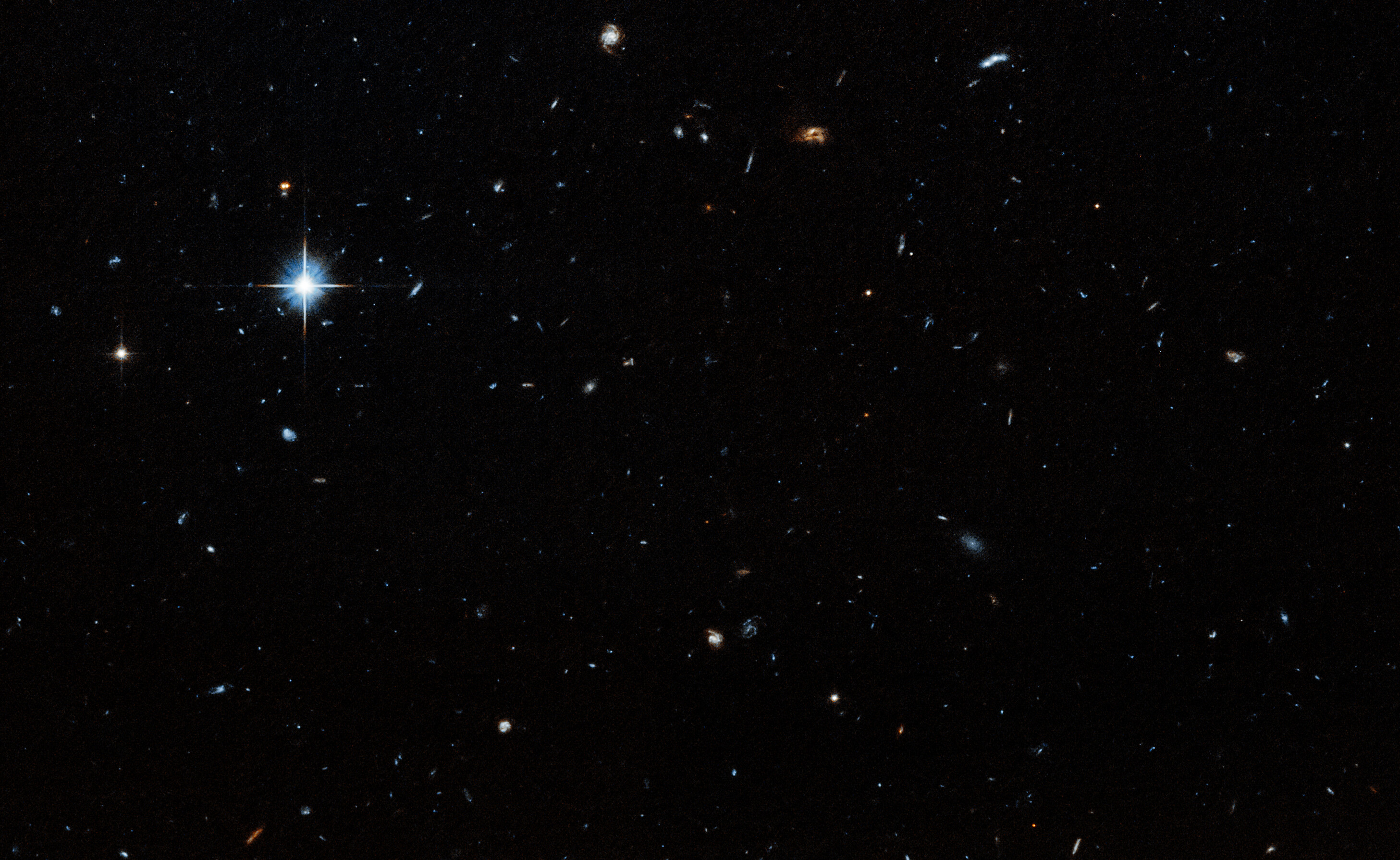 A dark field with stars and galaxies of various sizes speckled throughout the image. A particularly bright star is visible in the upper left region of the image.