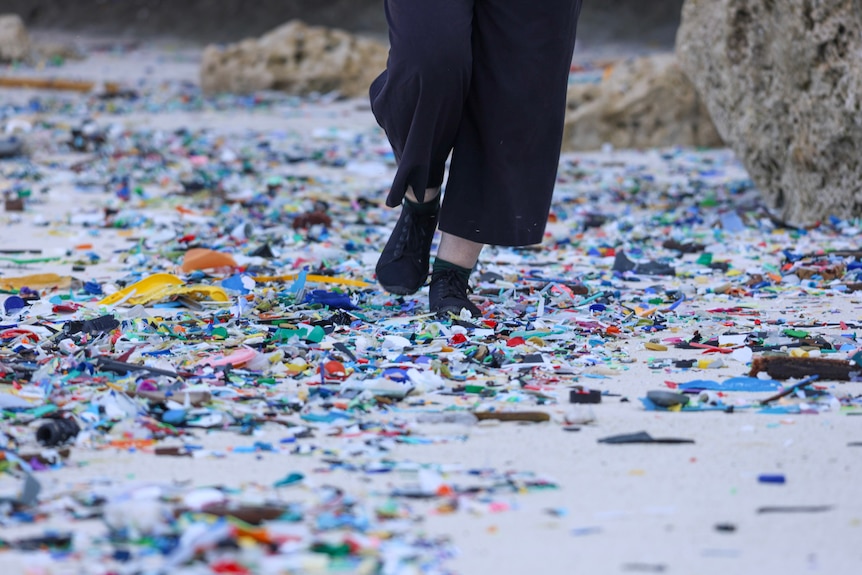 A photo of someone's legs wearing black pants and shoes walking along a thick layer of coloured plastics on a sandy beach.