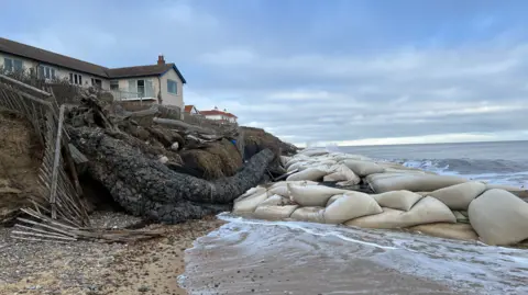 Richard Daniel/BBC A home on the edge of an eroding cliff in Thorpeness. There is a big cluster of geobag sea defences on the beach in front of it. The tide is washing in.