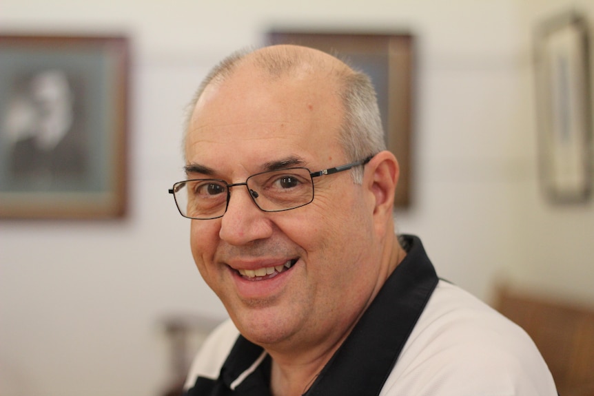 A bald man with black glasses and a black and white polo shirt smiles at the camera. 