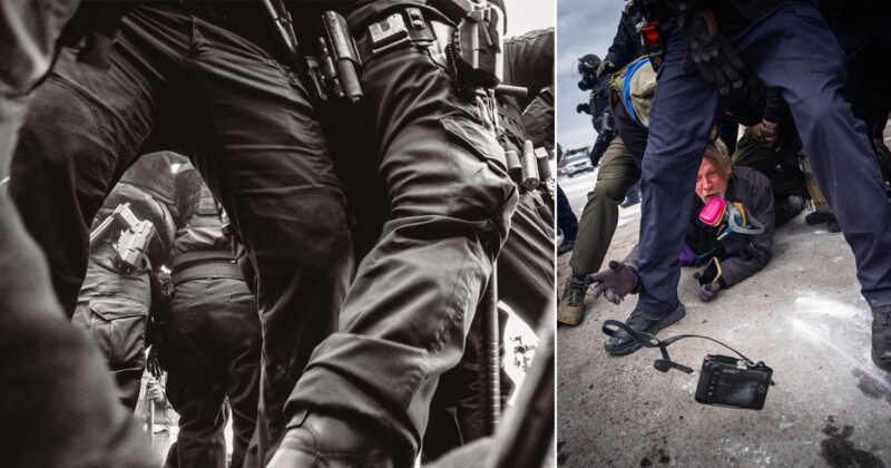 A man wearing a gas mask crouches on the ground amid armed police officers in tactical gear; a camera lies on the pavement nearby. The scene appears tense and chaotic.