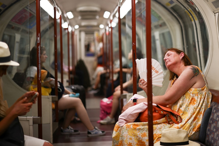 A woman looking very hot and sweaty fans herself with a paper fan while sitting on the London tube
