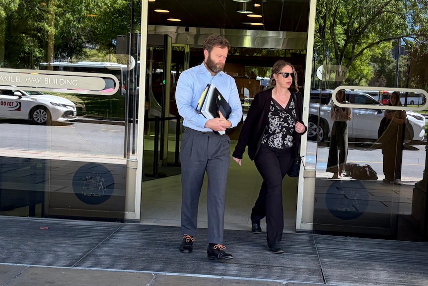 A woman and a man walk through glass doors, leaving a building